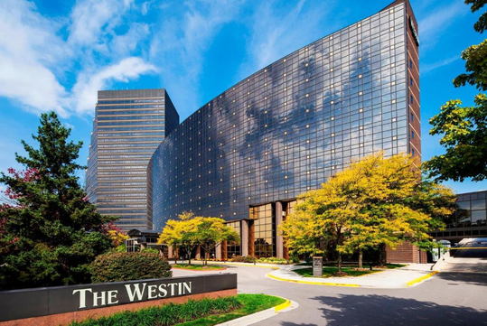 Exterior view of The Westin hotel featuring a modern glass building, trees, and a clear blue sky.