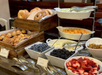 A buffet table with bagels, pastries, blueberries, blackberries, strawberries, melon, and cucumbers in bowls, with serving utensils.