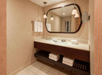 Bathroom with a large mirror, sink, and countertop. Towels are neatly arranged on the shelf below and hanging on a rack. Ceiling lights illuminate the area.