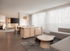 Modern living and dining area with a beige sofa, round wooden coffee tables, dining table with chairs, and a large TV. Large windows with sheer curtains allow natural light.