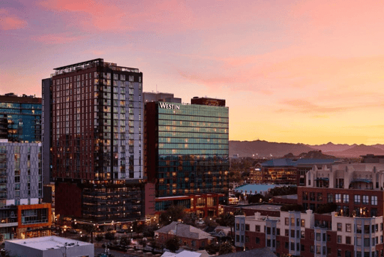 A cityscape at sunset features modern buildings, including a Westin hotel, with mountains in the background under a colorful sky.