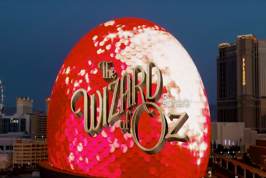 A large dome-shaped building displays a red, sequin-like light pattern with the text "The Wizard of Oz at Sphere" overlaid; city buildings are visible in the background.