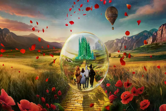 Three characters walk on a yellow brick road toward a green city inside a bubble, surrounded by fields, flowers, mountains, and a hot air balloon in the sky.