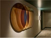 Abstract wall art composed of overlapping circular shapes in shades of brown, gold, and blue, displayed in a dimly lit hallway with patterned carpeting and a wall-mounted light.
