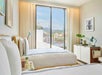 Bright hotel room with two double beds, a small desk, and a large window showing a cityscape. White bedding and neutral decor create a clean atmosphere.