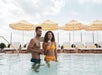 A man and woman stand in a pool holding drinks. Behind them are yellow and white striped umbrellas and lounge chairs.