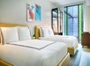 A hotel room features two neatly made double beds, framed artwork on the wall, a glass wardrobe with clothes, and a large window with a garden view.