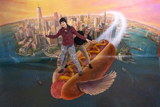 Two people pose as if riding a giant hot dog flying over a painted cityscape backdrop resembling New York City.