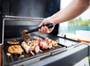 Person grilling skewers and meat on an outdoor gas grill using tongs, with visible smoke and a plastic container nearby.