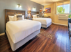 Hotel room with two large beds, wooden floors, wall art, and a window with a view of trees. Decor includes patterned pillows, bedside lighting, and a small table with a chair.
