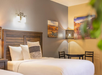 Hotel room with two beds, wooden headboards, wall art, a table with two chairs, and a lamp.