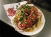 A plate of tacos topped with chopped onions, cilantro, diced tomatoes, and pickled red onions sits on a paper plate.