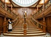 A woman in a maid costume stands at the bottom of an ornate wooden staircase with decorative railings and a statue at the center.
