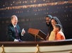 A man in a suit performs a card trick for a smiling family of three seated behind a wooden podium, with a ship backdrop in the background.