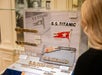 A woman views a museum display case containing S.S. Titanic nameplates and White Star Line flags, with informational plaques visible.