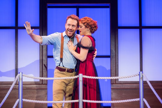 A man in suspenders gestures and smiles while a woman in a red dress stands beside him on a stage set with a rope railing and a blue-lit background.