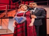 A woman in a red dress with a large blue heart stands with a man in a tuxedo on a stage set with stairs and ropes.