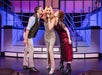 Three performers on stage: a man on the left, a woman in a glittering dress singing in the center, and a woman in a red dress on the right. The background has pink and blue lighting.