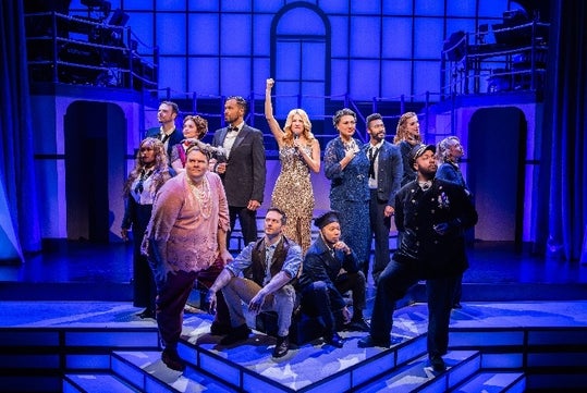 The cast of Titanique on Broadway posing on stage with purple light shining down on them.