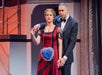 A woman in a red dress with a large blue heart necklace stands beside a man in a black tuxedo; both face forward on a stage set.