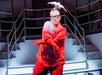 A person in red gloves and clothing poses dramatically on a white staircase stage, wearing glasses and a headpiece with a white bird figure.