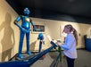A young woman interacts with an exhibit featuring a blue humanoid robot in a science or technology museum.