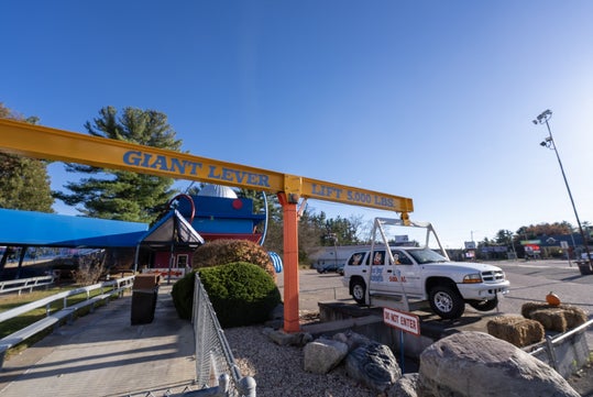 A large yellow lever labeled "Giant Lever Lift 5000 lbs" overhangs a parking lot with a white truck beneath it; a building and blue awnings are in the background.