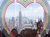 A tower viewer in front of a rainbow heart on the glass at the Top of the Rock Observation Deck with New York City in the background.