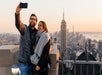 A couple in winter clothes standing taking a selfie with the Empire State Building in the background at sunset.