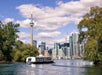 A boat travels on water with the Toronto skyline and CN Tower in the background, framed by trees under a partly cloudy sky.