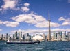 A boat travels on a body of water in front of the Toronto skyline, featuring the CN Tower and various modern high-rise buildings under a partly cloudy sky.