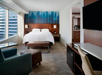 Modern hotel room with a king bed, brown headboard, blue artwork, armchair, ottoman, large window, desk, and TV. Beige carpet and neutral decor create a sleek and inviting atmosphere.