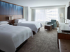 Hotel room with two double beds, a large window, armchair, ottoman, desk, and wall-mounted TV. Neutral decor with blue and brown accents.