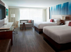 Hotel room with two double beds, a sofa, round table, chair, wall-mounted TV, and large window with curtains. The decor features a neutral color palette and wood flooring.