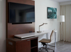 Minimalist hotel room corner with a wall-mounted TV, white desk with a lamp and chair, and a floor lamp. Decor includes a framed abstract art piece on the wall.