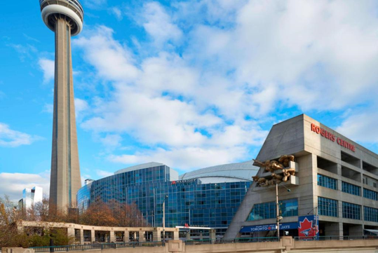 The image shows the CN Tower beside the Rogers Centre with its sign visible. The sky is partly cloudy, and some trees are in the foreground.