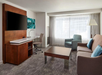 A modern hotel room features a sofa, coffee table, armchair, large TV, and desk with a chair. The room has a large window with curtains, providing natural light.