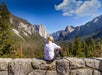 A hiker in a white shirt sitting on a rock wall over looking Yosemite National Park on a sunny day with clouds in the sky.