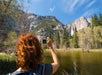 A hiker with bright red hair taking a picture of a mountain with a lake in front of it on a sunny day in Yosemite National Park.