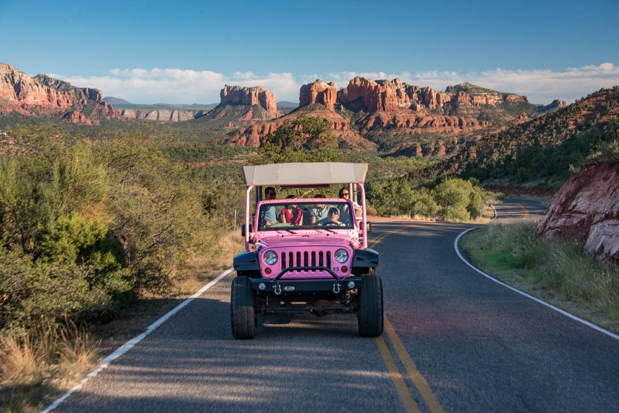 A pink jeep with passengers drives on a winding road through a desert landscape with red rock formations and sparse vegetation under a clear sky.
