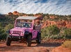 A pink off-road tour jeep with passengers drives on a dirt path through a desert landscape with red rock formations and scattered vegetation.