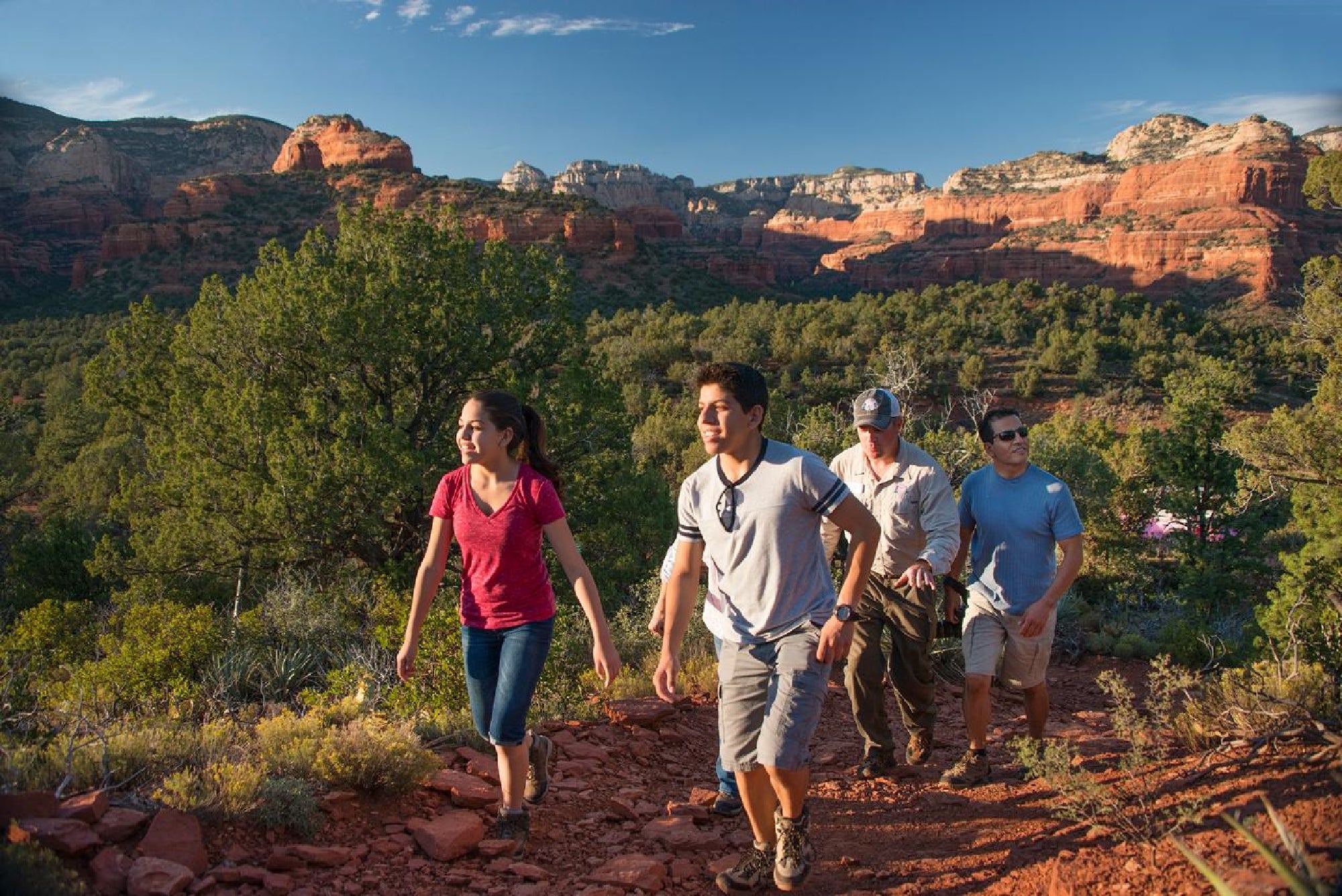 Four people hike on a rocky trail surrounded by trees, with red rock formations and a blue sky in the background.