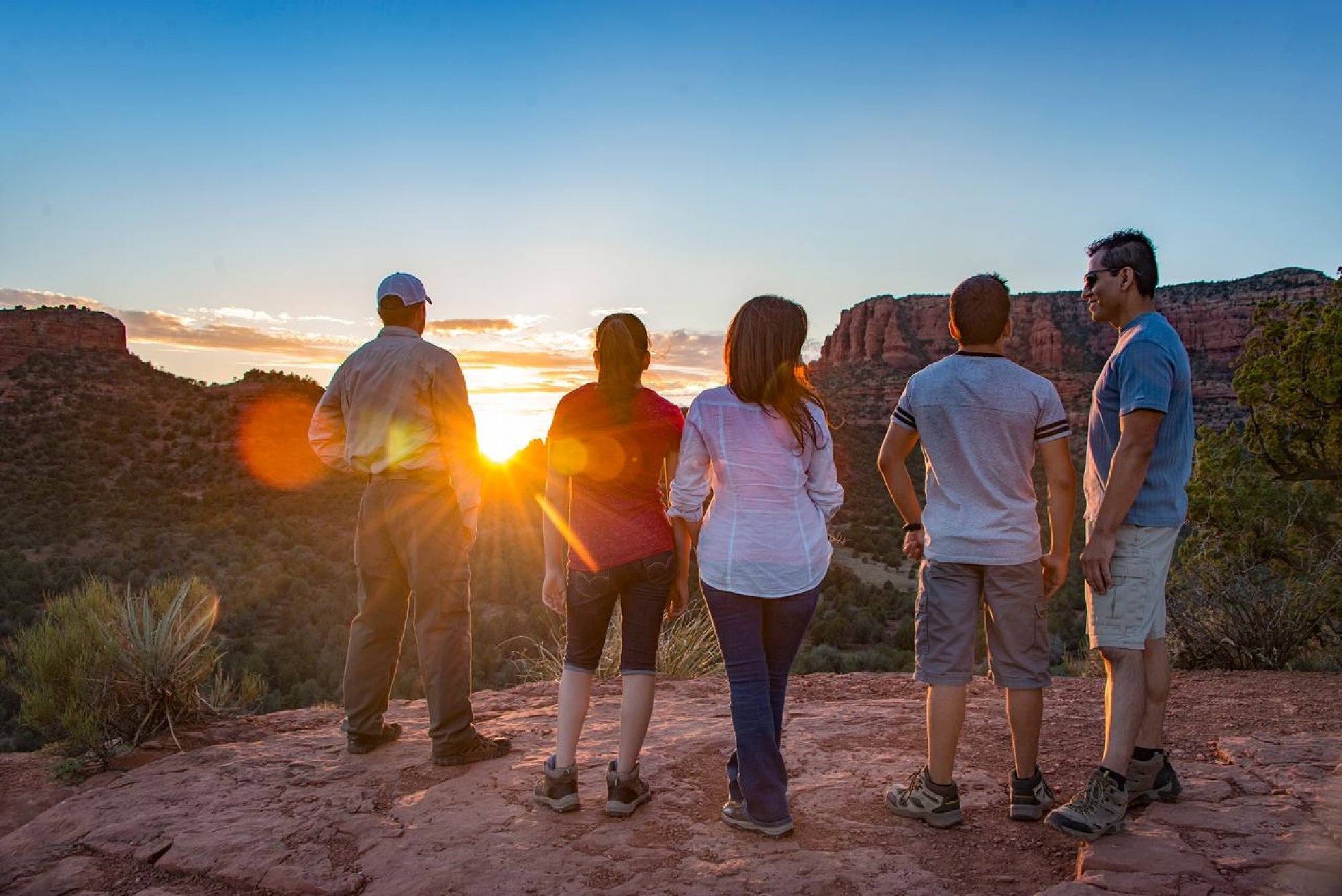 Five people stand on a rocky ledge, facing a sunset over a canyon landscape with vegetation and red rock formations.