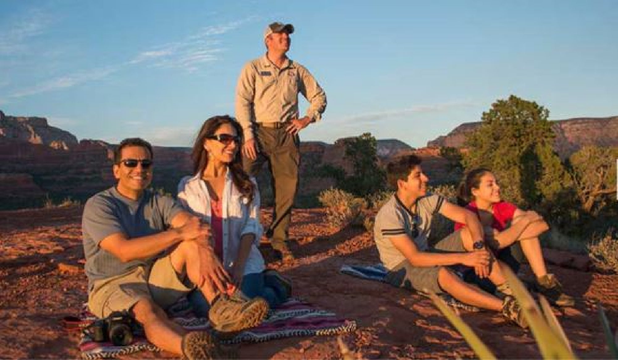 Four people sit on a rocky landscape with a guide standing behind them, all looking towards the distance in an outdoor setting.