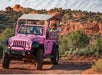 A bright pink Jeep with a canopy drives along a dirt road in a desert landscape with red rock formations and green shrubs.