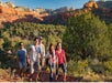 Five people hike along a rocky trail surrounded by green trees, with red rock formations and a blue sky in the background.