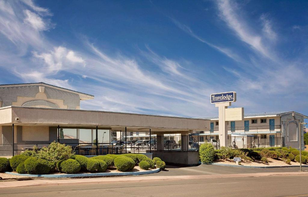 Roadside motel with a "Travelodge" sign, featuring a one-story building, manicured shrubs, clear blue sky, and light cloud formations.
