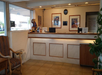 Hotel reception desk with wooden accents and framed art on the wall. To the left, a striped chair is placed near a window, and a potted plant partially visible on the right.