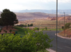 A quiet road curves through a landscape featuring green lawns, trees, and distant reddish hills under a cloudy sky. A sign by the road indicates "Full Breakfast Quality Inn".