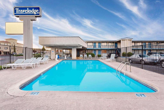 Outdoor swimming pool at a Travelodge hotel, surrounded by lounge chairs and a metal fence, with the hotel building and parking lot visible in the background.